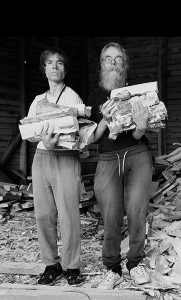 Mustavalkoinen dokumentaarinen muotokuva: kaksi henkilöä kantaa halkoja vanhassa vajassa. Kuvaajana Johanna Kare.